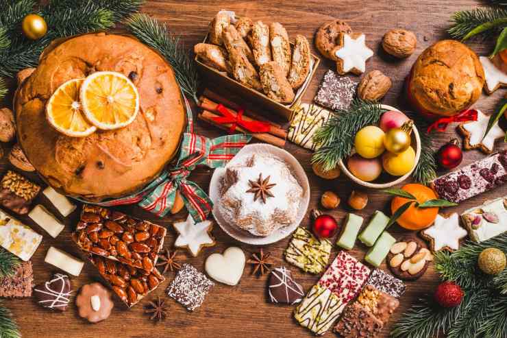 Un tavolo pieno di dolci natalizi assortiti, tra cui panettone, pandoro, biscotti decorati, torrone, frutta secca e cioccolatini. Al centro, un pandoro decorato con anice stellato e un panettone guarnito con fette di arancia, circondati da decorazioni natalizie come rami di pino e palline colorate.